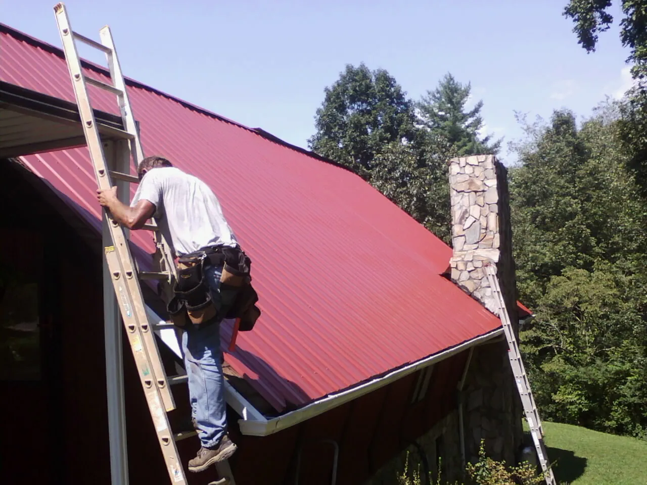 Roofer on a ladder beside a red metal roof and stone chimney — Soffit & Fascia Repair by Roofing Brothers in Evanston